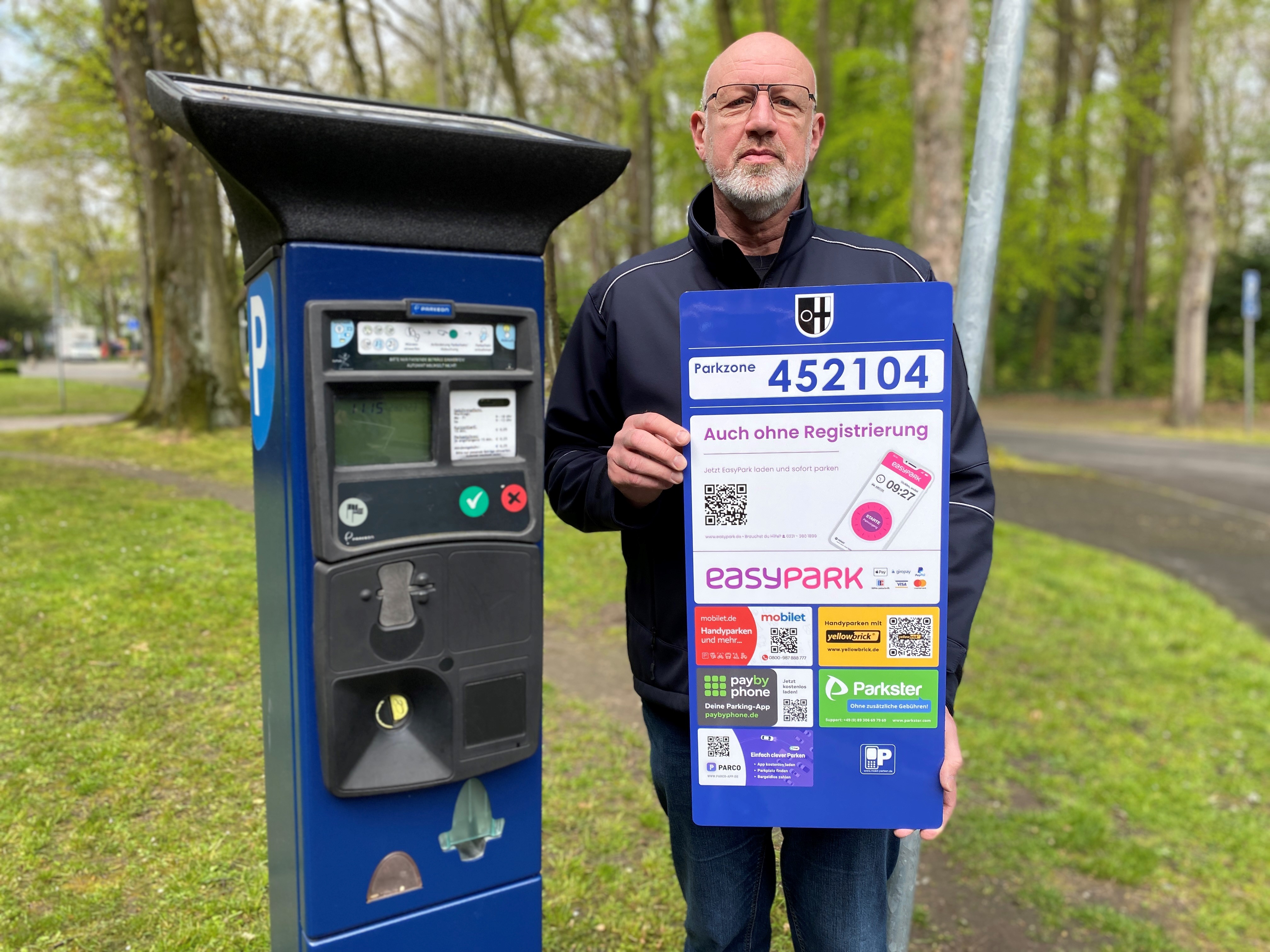 Ralf Feldhaus, Fachdienstleiter Verkehrsangelegenheiten, präsentiert am Parkautomat vor dem Rathaus ein Hinweisschild für das Handyparken.
