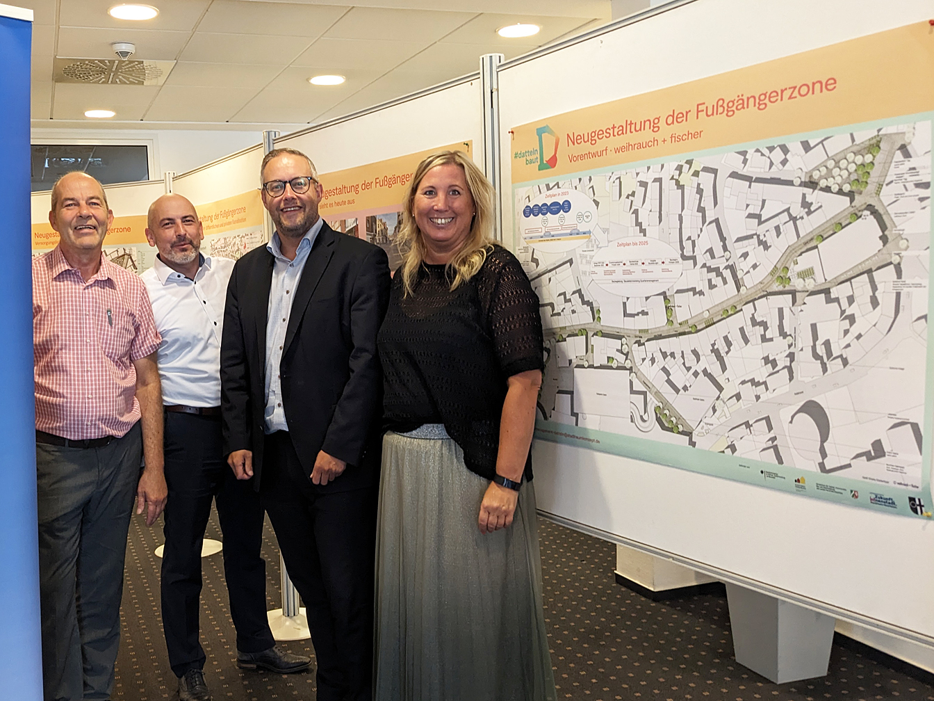 Vier Personen stehen in den Räumen der Volksbank vor den Plänen zur Umgestaltung der Innenstadt