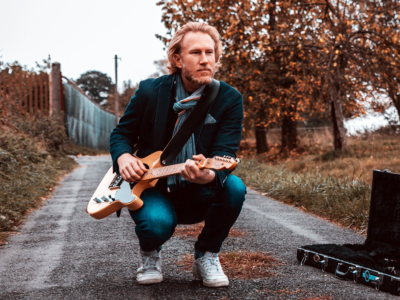Musiker und Gitarrist Phil Solo, der mit einer E-Gitarre in der Hand auf einer Straße kniet; im Hintergrund sind Bäume und ein Zaun zu sehen