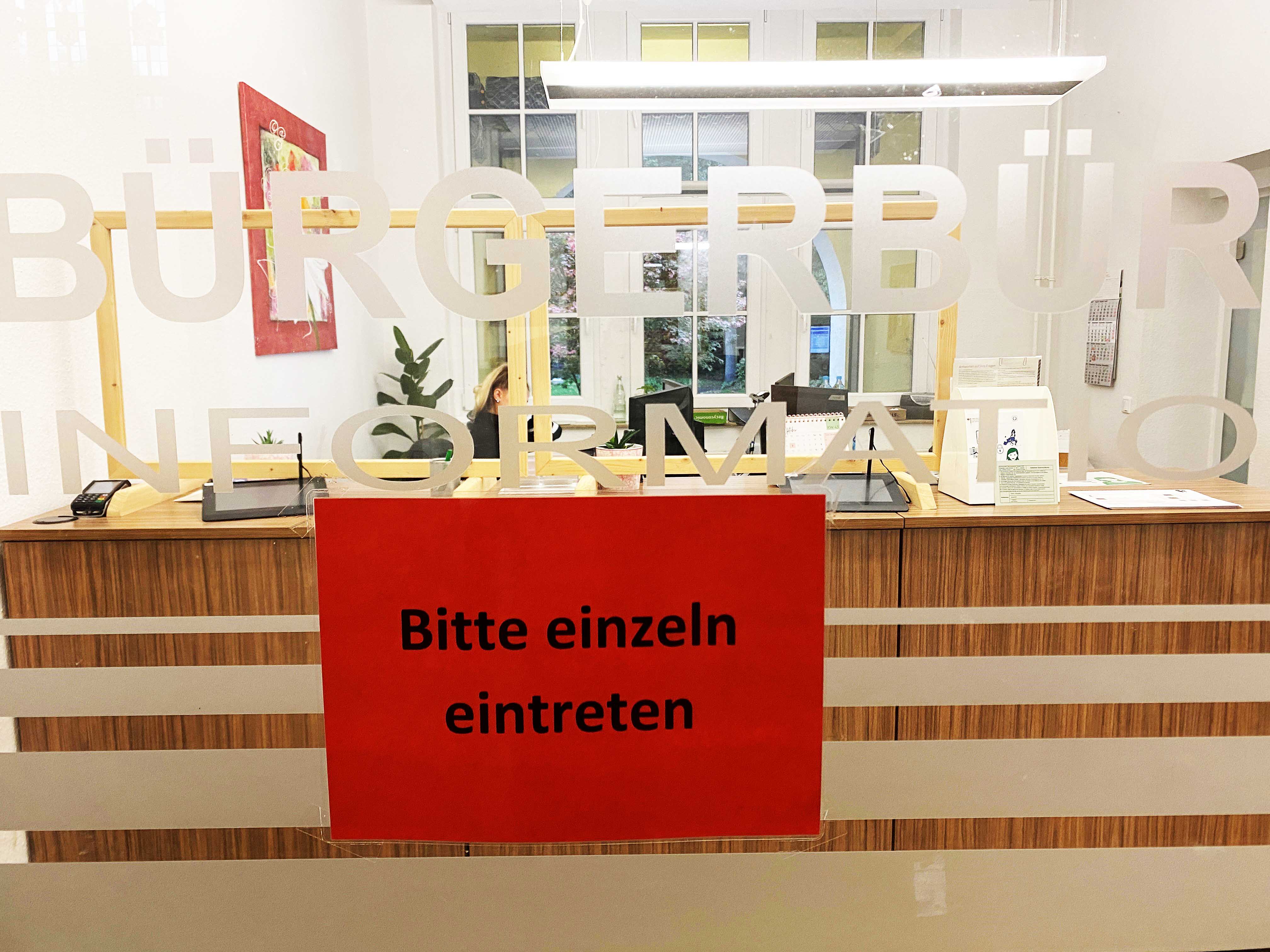 Eingang zum Bürgerbüro im Rathaus; auf der Glastür ist ein Schild mit der Aufschrift "Bitte einzeln eintreten" zu sehen, darüber steht "Bürgerbüro" und "Information"; im Hintergrund ist die Infotheke des Bürgerbüros.