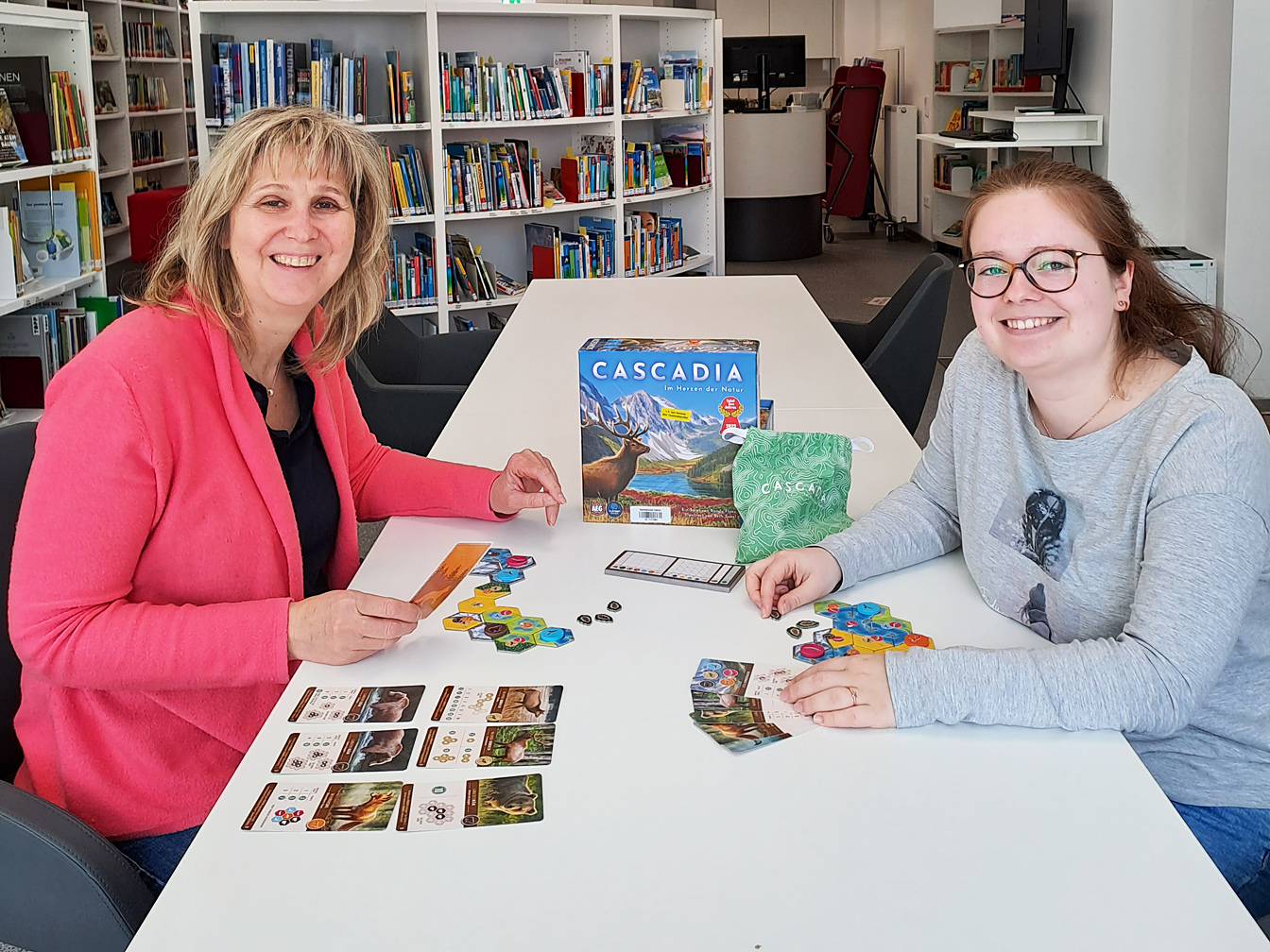 Sybille Schumacher (links) und Kiara Bender sitzen am  Tisch und spielen ein Gesellschaftsspiel in der Stadtbücherei