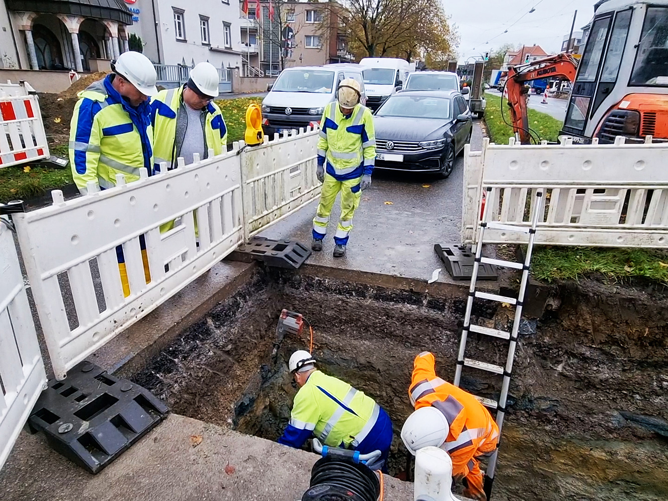 Bauarbeiter stehen in einer Baugrube, andere stehen außerhalb der Baustellenabsperrung; im Hintergrund stehen Autos, rechts ist ein Bagger zu sehen und links die Moschee.