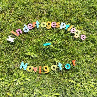 Das Wort "Kindertagespflege-Navigator" ist mit einzelnen bunten Buchstaben auf einem Rasen gelegt worden.