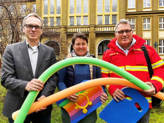 Das Bild zeigt von links Bürgermeister André Dora, Raphaela Tewes (Leiterin Schul- und Sportverwaltung) und Joachim Haas (Beauftragter Rettungsschwimmen beim DLRG), die vor dem Rathaus stehen und für das Foto Schwimmutensilien wie Schwimmnudeln in der Hand halten.