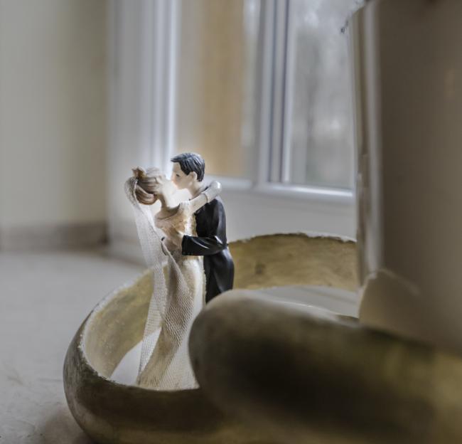 Das Bild zeigt zwei kleine Figuren, die ein Brautpaar darstellen sollen, die sich in einem überdimensionalen Ehering umarmen