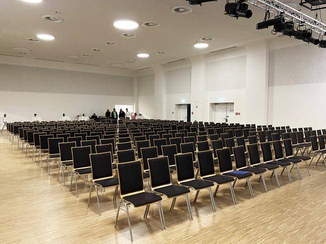 Stühle, die in Reihe stehen, in der Stadthalle. Im Hintergrund stehen fünf Personen.
