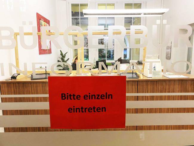 Eingang zum Bürgerbüro im Rathaus; auf der Glastür ist ein Schild mit der Aufschrift "Bitte einzeln eintreten" zu sehen, darüber steht "Bürgerbüro" und "Information"; im Hintergrund ist die Infotheke des Bürgerbüros.