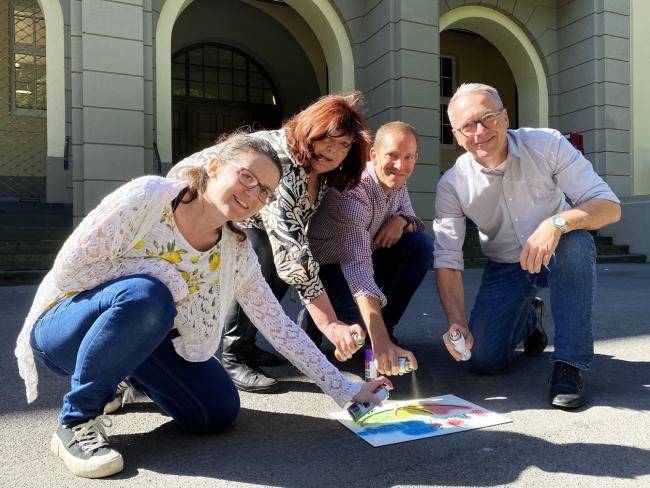 Die städtischen Mitarbeiter*innen Patricia Magiera, Susanne Sassor und Julian Sauck (v. l.) sprühen zusammen mit Bürgermeister André Dora (r.) das Motto der Demokratiewoche „Viele gemeinsam für alle“ auf den Rathausvorplatz.