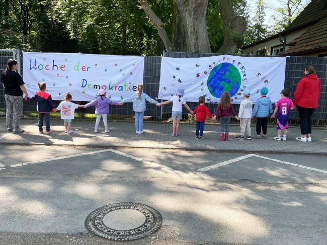 Kinder und Erzieher*innen der Dependance des Städtischen Bewegungskindergarten an der Berliner Straße stehen vor Plakaten zur Woche der Demokratie