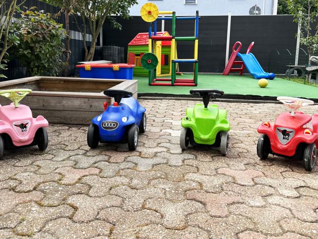 Vier Rutschautos in Reihe vor einem Sandkasten, im Hintergrund Spielgeräte